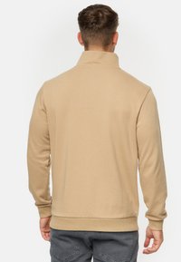 Sweatshirt beige zippé avec un col haut, des poignets et un ourlet côtelés, présentant une texture lisse et une silhouette ajustée sans embellissements visibles.