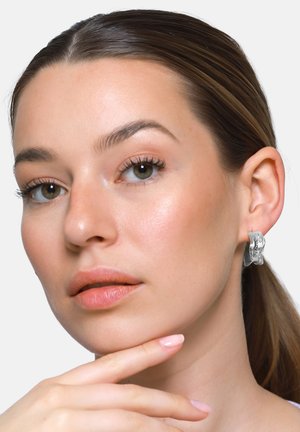 Donna con pelle chiara e capelli castani che indossa orecchini a cerchio argentati lucidi e testurizzati, appoggiando il mento sulla mano con unghie rosa chiaro.