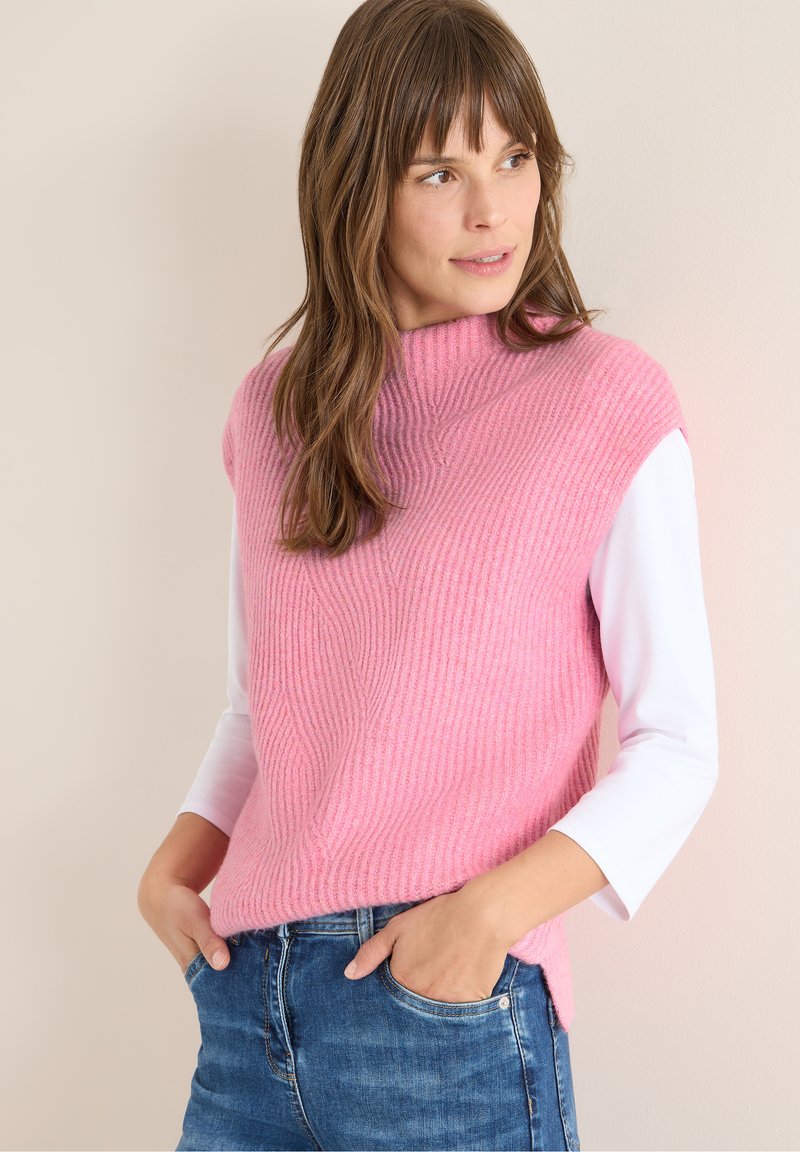 Vaaleanpunainen ribattua neulosta oleva hihaton toppi, jossa on korkea kaula-aukko, yhdistettynä valkoiseen pitkähihaiseen paitaan alla. Sisältää sivuhalkiot ja rentoa istuvuutta.