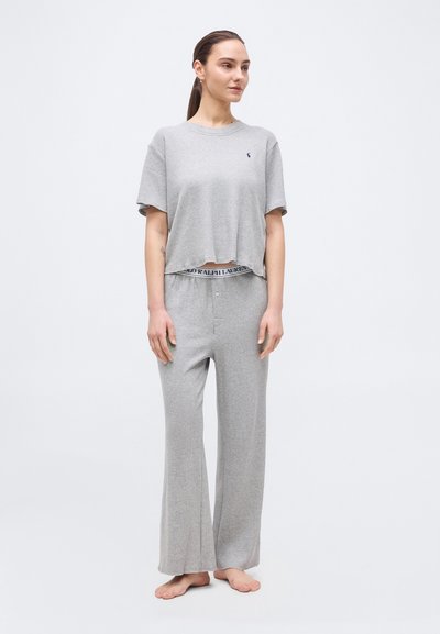 Νεαρή γυναίκα όρθια ξυπόλητη, φορώντας άνετο ανοιχτογκρί κοντομάνικο τοπ και παντελόνι φαρδιάς γραμμής, μπροστά από απλό φόντο.