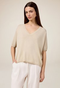 Beige V-Ausschnitt Stricktop mit kurzen Ärmeln, in lockerer Passform und mit geripptem Saum. Kombiniert mit weißen, plissierten Hosen.