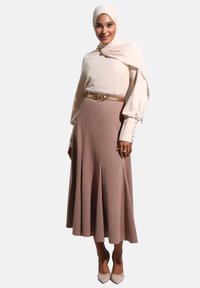 Blouse beige clair à manches longues, jupe rose pâle taille haute, ceinturée avec des accessoires dorés, et escarpins nude à bout pointu. Textures lisses tout au long.
