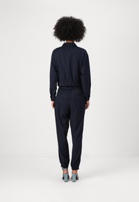 Tuta blu navy con righe a spina di pesce, cintura e polsini elastici. Tessuto morbido e liscio; scarpe coordinate con tacco alto in azzurro chiaro.