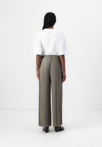 Femme avec des cheveux tressés portant un haut blanc à manches courtes, un pantalon large plissé vert olive et des chaussures à talons noires, faisant face à un mur blanc.