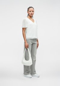 Γυναίκα με πλεξούδες φοράει λευκή διάφανη μπλούζα με κοντά μανίκια, γκρι τζιν παντελόνι ίσιου στριφώματος, λευκά αθλητικά παπούτσια και κρατά λευκή τσάντα χειρός με κοψίματα.