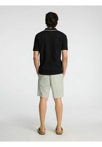 Črna polo majica s kontrastnim ovratnikom, v kombinaciji z svetlimi kratkimi hlačami z nizkimi črtami. Model stoji bos z sproščenim držanjem.