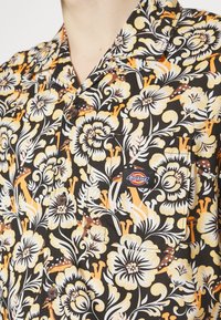Gros plan sur une chemise noire boutonnée avec un motif floral orange et beige, avec une poche avant portant une étiquette de la marque Dickies.