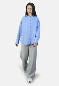 Camisa de botones azul claro con cuello, mangas largas y corte holgado; combinada con pantalones grises de pierna ancha y zapatillas deportivas blancas.