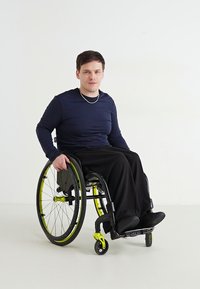 Χειροκίνητο αναπηρικό καρότσι με μαύρο σκελετό, κίτρινες λεπτομέρειες και σταθερές ρόδες. Ο χρήστης φοράει μπλε μακρυμάνικο πουκάμισο και μαύρο παντελόνι, καθισμένος άνετα.