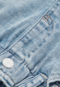 Close-up af lyseblåt denimstof med detaljeret syning og en sølv metal knap indgraveret med tekst.