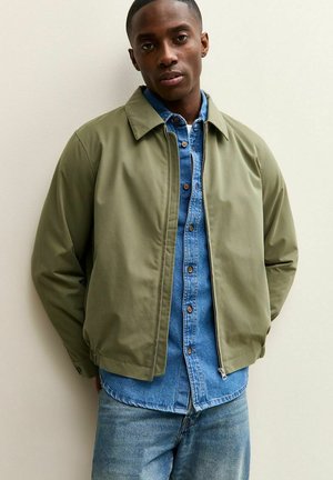 Olijfgroene bomberjack met een ritssluiting aan de voorkant, daaronder een denim blouse met knopen en lichtblauwe spijkerbroek. Soepele, lichte stof.