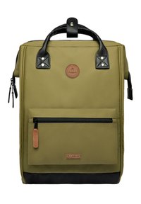 Olive green backpack with a textured black bottom, top handle, front zip pocket, and circular brand patch. Smooth, durable material.