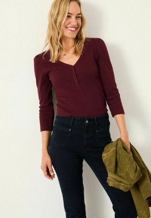 Burgundy long-sleeve top with a V-neck and button details, paired with dark denim jeans. Green suede jacket held in one hand.