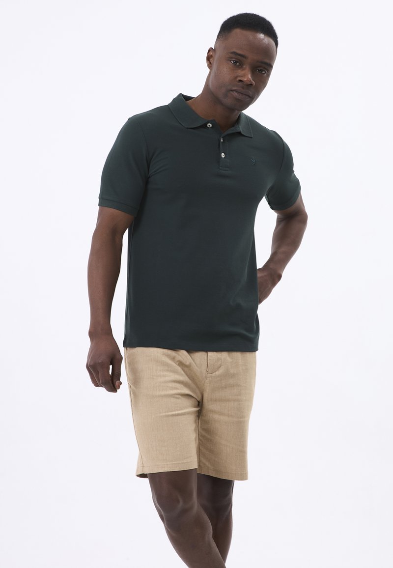 Donkergroen polo shirt met kraag en drie knopen, gecombineerd met lichtbruine shorts, weergegeven tegen een eenvoudige witte achtergrond.