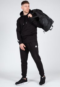 Felpa nera con tasca frontale, pantaloni della tuta neri coordinati e scarpe da ginnastica nere. Logo bianco notevole su felpa e pantaloni. Il materiale sembra morbido.