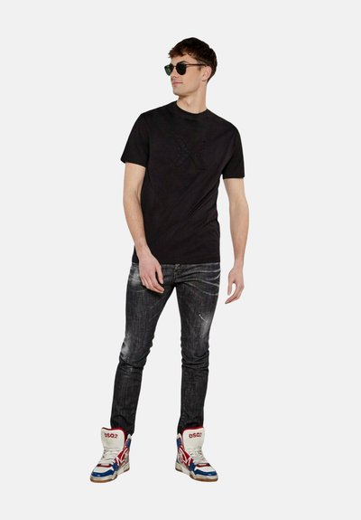 T-shirt nera in cotone con grafica in rilievo, abbinata a jeans in denim scuro con effetto usurato e sneakers alte con accenti rosso, bianco e blu.