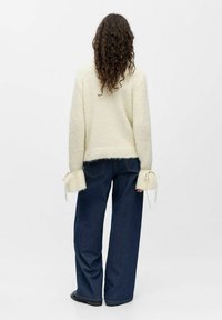 Suéter de punto crema con mangas anchas que presentan lazos en los puños, combinado con unos pantalones de mezclilla de pierna ancha en azul oscuro. Textura suave y corte relajado.