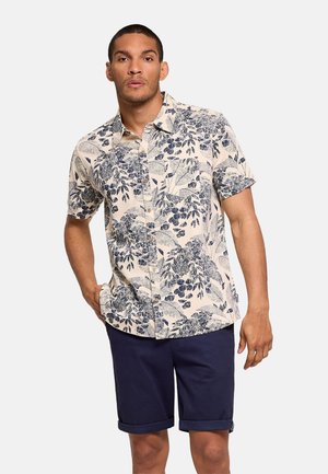 Hombre con una camisa beige de manga corta con botones y diseños de hojas azules y pantalones cortos azul marino enrollados, de pie con una mano en el bolsillo.