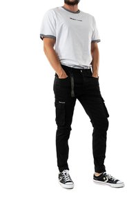 Pantalones cargo negros con múltiples bolsillos, combinados con una camiseta blanca de cuello redondo con ribetes negros y texto de logo, llevada con zapatillas negras.