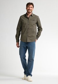 Verkeerde button-up shirt in donker groen en beige, fitted ontwerp met een borstzak, gecombineerd met blauwe jeans en witte sneakers.