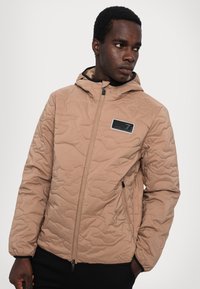 Beige quiltet jakke med hætte, der har en frontlynlås, sorte detaljer og et mærket patch på brystet. Glat tekstur, letvægtsdesign.