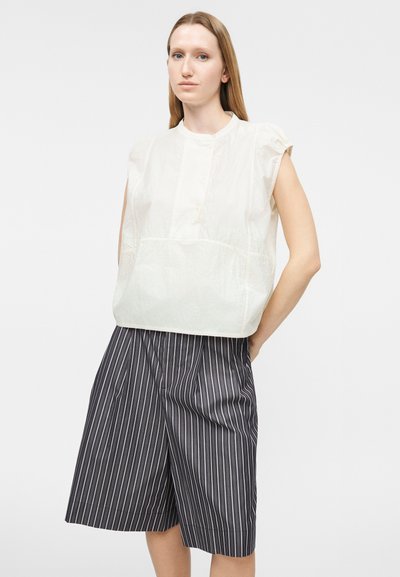 Γυναίκα με ίσια ανοιχτό καστανά μαλλιά φοράει αμάνικο λευκό πουκάμισο και φαρδιά μαύρα σορτς με λευκές κάθετες ρίγες, στέκεται με τα χέρια πίσω από την πλάτη.