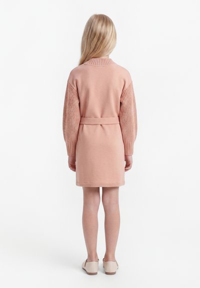 Niño con cabello largo y rubio de espaldas, vestido con un vestido rosa con cinturón, mangas de punto texturizadas y zapatos planos beige.