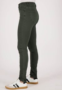 Skinny jeans in olijfgroen, gemaakt van rekbaar materiaal, met achterzakken en een tailleband met riemlussen, gedragen met witte sneakers.