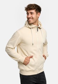 Crèmekleurige hoodie met rits van een zachte stof, voorzien van twee zakken aan de voorkant, een capuchon met trekkoord en geribbelde boorden. Ontworpen voor comfort en warmte.