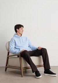 Light blue hooded sweatshirt, dark brown pants, and black sneakers. Model seated on a wooden chair with a beige cushion. Minimalist background.