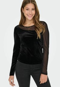 Mladá žena v černém sametovém topu s průhlednými dlouhými rukávy a černých kalhotách, usmívající se s volně vlnitými vlasy a zlatými šperky.