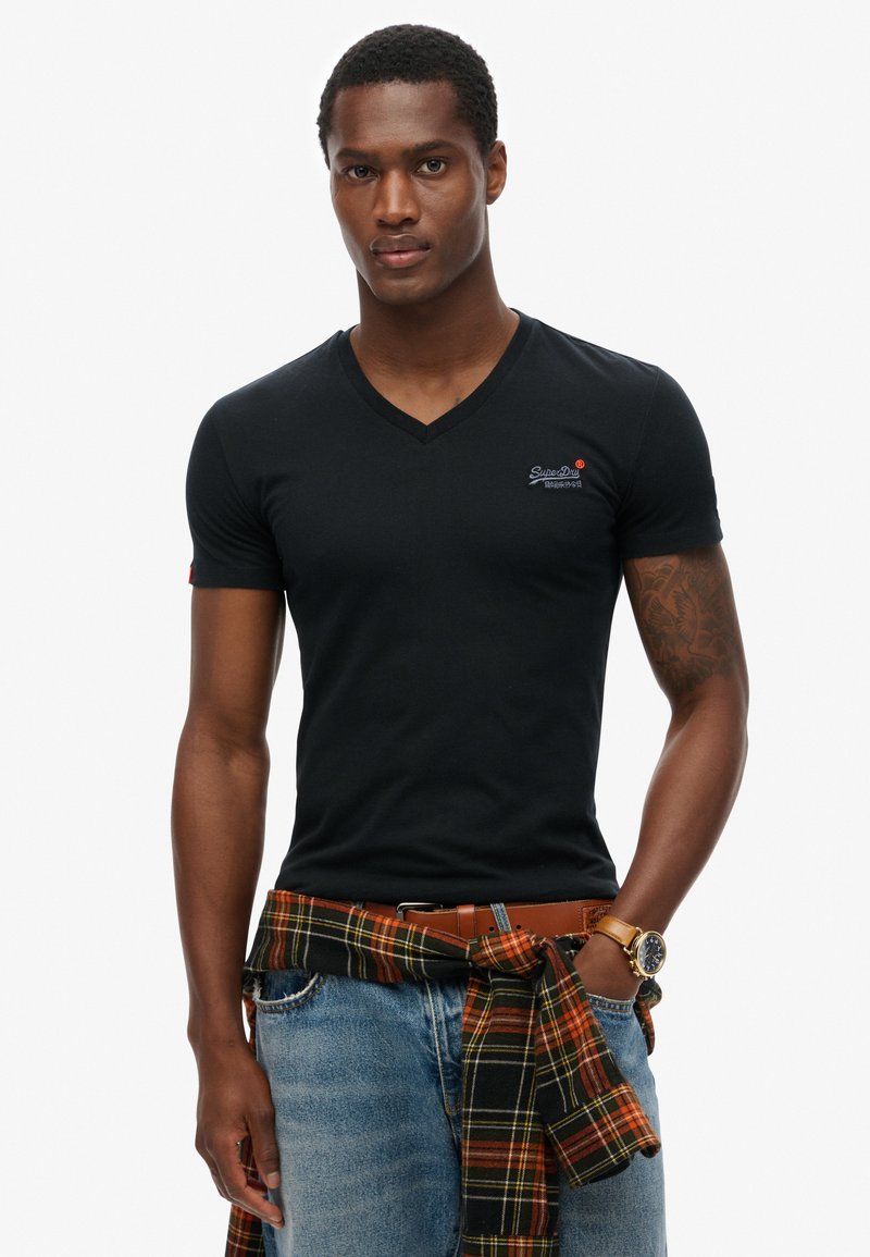 Camiseta negra de cuello en V con mangas cortas, con un logo en el pecho. Combinada con unos jeans azules y una camisa de cuadros atada en la cintura.