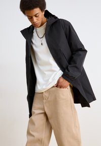 Zwarte waterdichte jas met capuchon en elastische manchetten, gedragen over een wit T-shirt en beige broek. Heeft een minimalistisch ontwerp en zakken.
