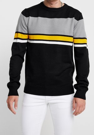 Mężczyzna w czarnym swetrze z poziomymi paskami w kolorach szarym, żółtym, białym i czarnym na klatce piersiowej i górnych rękawach, w połączeniu z białymi spodniami.
