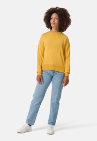 Maglione giallo a maglia con scollo rotondo, taglio rilassato e orlo a coste, abbinato a jeans azzurri chiari e sneakers bianche.