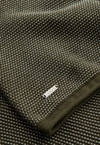 Gros plan d'un tissu tricoté texturé vert foncé et blanc avec une petite étiquette métallique portant la mention "ombrec" près du bord du vêtement plié.