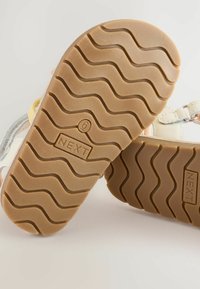 Jasne dziecięce buty z gumową podeszwą, z teksturowanym, falistym wzorem oraz logotypem "NEXT" na spodzie.