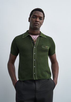 Hombre con camisa de botones de manga corta de color verde oscuro texturizada con costuras en contraste y pantalones oscuros, de pie con las manos en los bolsillos frente a un fondo gris.