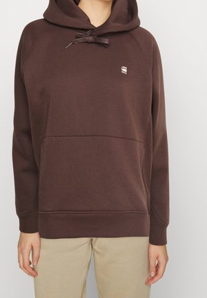 Osoba ubrana w brązową bluzę z kapturem ze ściągaczami i małym logo na piersi, w połączeniu z beżowymi spodniami, stojąca na jednolitym tle.