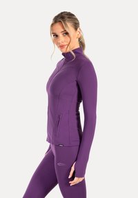 Chaqueta ajustada de color morado con cierre de cremallera, alto cuello, bolsillos laterales con cremallera y mangas alargadas. Material suave y elástico con costuras planas.
