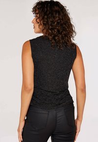 Ärmelloses schwarzes Top mit dezentem metallischem Schimmer, strukturiertem Stoff und gerafftem Rückendesign, kombiniert mit enganliegenden schwarzen Hosen.