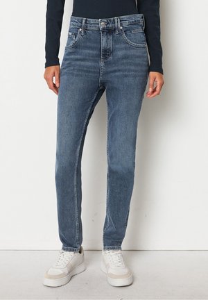 Vrouw die hoge taille strakke blauwe jeans en witte sneakers draagt, staand tegen een effen muur met handen ontspannen naast het lichaam.