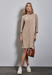 Robe en tricot beige avec une texture subtile, manches trois-quarts et coupe droite. Associée à des baskets blanches et un sac à main marron.
