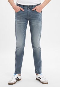 Svetlomodré džíny so slim fit, vybavené gombíkovým zapínaním a piatimi vreckami; nosené s bielymi teniskami s čiernymi pruhmi.
