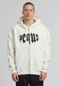 Witte zip-up hoodie met een grote zwarte grafische print op de borst. Voorzien van een trekkoordkap en zijzakken. Zachte stoftextuur.