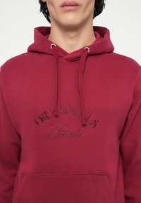 Burgundia kapuutsiga jakk, millel on paelasüsteem, tikitud "Originals Studio" logo, esitasku ja pehme puuvilla segu tekstuur.