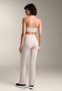 Beige sportmelltartó, amelynek hátul strappy dizájnja van, magas derekú, testhezálló bézs trapéznadrággal párosítva, feketemellényű sportcipővel kiegészítve.