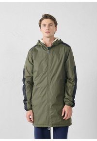 Groene parka-jas met een capuchon, zwarte mouwen, ritssluiting, bontgevoerde kraag en elastische manchetten. Voorzien van voorzakken en een tunnelkoord aan de zoom.