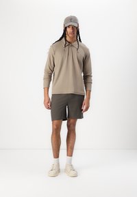 Haut à capuche beige à manches longues avec un col, associé à un short vert foncé et des baskets crème. Il présente des manches froncées et une coupe décontractée.