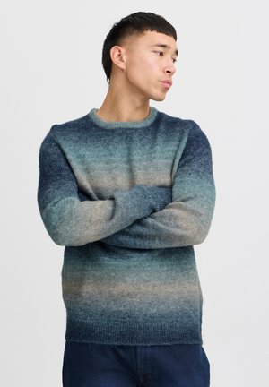 Suéter de punto con un degradado de azul profundo a beige suave, con un cuello redondo y una tela texturizada. Se lleva con los brazos cruzados.